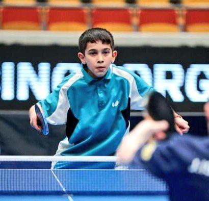A young boy with short dark hair wearing a light blue and white collared sports shirt and shorts stands on an indoor table tennis court holding a paddle in his right hand as he swings to hit a ball over the net toward the opposite side with a black background banner displaying text behind him and empty spectator seats in the blurred background.
