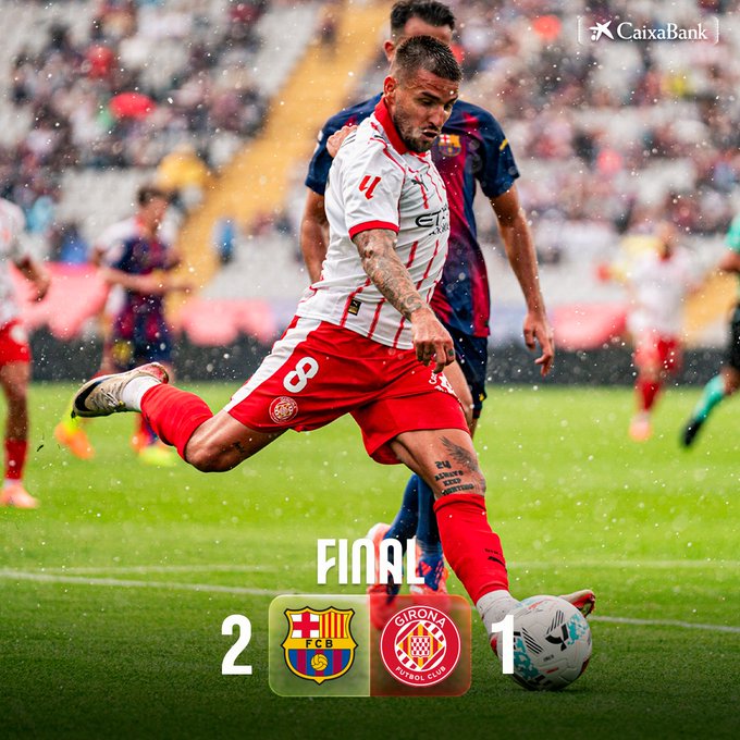 A soccer player in a white and red Girona FC jersey number 8 with tattoos on his arms and legs kicks a ball towards the goal during a match against Barcelona players in blue and red jerseys on a green field in a stadium with spectators and Caixabank sponsor logos visible. The score overlay shows Barcelona 2 Girona 1 final.