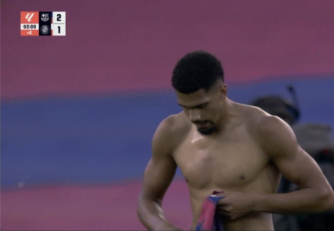 A screenshot from a football match broadcast shows a Black male player with short hair and a mustache standing shirtless on the field, holding a red towel over his shoulder with his right hand while his left arm is extended. He appears sweaty and focused, wearing blue shorts. The background includes a purple stadium seating area with blurred spectators. The scoreboard overlay in the top left displays UEFA branding, a score of 1-1, and time 90+9 minutes. The field has green grass with white lines.
