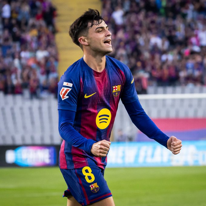 Young male athlete with dark hair wearing FC Barcelona blue and red jersey with number 8, sponsor patches including Spotify, shorts and socks in matching colors, standing on green soccer field with yellow stadium seating and blurred crowd in background, arms raised in celebration with open mouth expression.