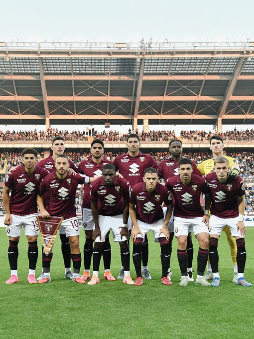 Eleven soccer players in maroon and white Torino FC uniforms with sponsor logos stand in two rows on a green field, the front row kneeling and holding a trophy shaped like a bull, the back row standing, all wearing cleats, in front of a large stadium with curved roof and seated crowd in the background under a clear sky.