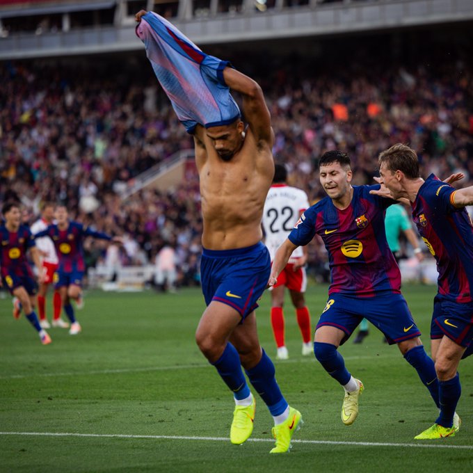 A shirtless male athlete with muscular build raises a blue jersey over his head in celebration on a green soccer field. Teammates in blue and red kits including numbers 9 and 22 surround him, some gesturing excitedly. A white-uniformed opponent player looks on. Stadium seating with blurred crowd and orange accents visible in background. Sponsor logos like Spotify on jerseys.