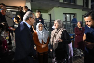 First image shows a group of people including men in suits and women in headscarves standing and talking outdoors near a green building with a fence in the background. Second image depicts several men in formal attire and vests with Turkish flag patches gathered in conversation outside a building. Third image features a diverse group of men women and a child posing near a white van and orange containers in an open area with buildings around. Fourth image captures individuals including a man in a suit and others near shelves stocked with toilet paper products inside a store with pink lighting.