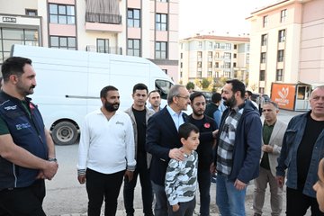 First image shows a group of people including men in suits and women in headscarves standing and talking outdoors near a green building with a fence in the background. Second image depicts several men in formal attire and vests with Turkish flag patches gathered in conversation outside a building. Third image features a diverse group of men women and a child posing near a white van and orange containers in an open area with buildings around. Fourth image captures individuals including a man in a suit and others near shelves stocked with toilet paper products inside a store with pink lighting.