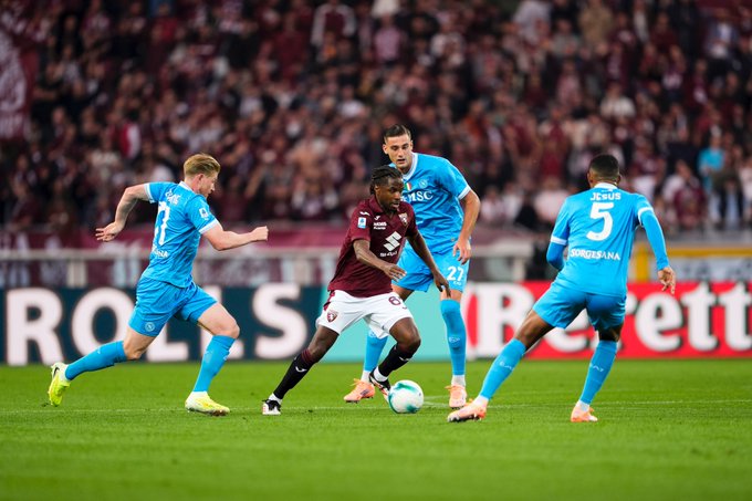 Four soccer players in action on a green field during a match. Two in blue kits with numbers 24 and 5, one in maroon kit with number 28 holding the ball, and one in blue kit with number 8 approaching. Stadium stands filled with red-shirted crowd in background. Sponsor board with Rolland Garros text visible. Players wear cleats and shorts.