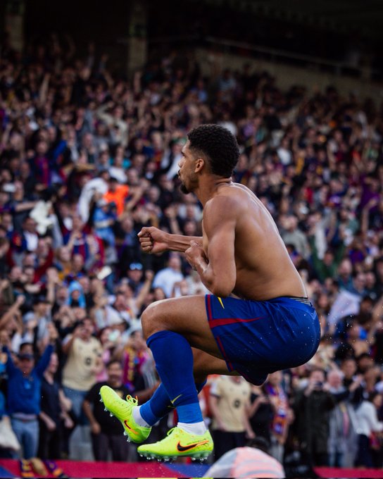 Shirtless male soccer player with short curly hair jumps triumphantly on a stadium field, arms raised in celebration, wearing blue shorts with red stripes, blue socks, and bright yellow cleats. Surrounding him are blurred crowds of fans in the stands, some waving arms and wearing orange clothing. The scene captures a moment of victory in a professional match for FC Barcelona.