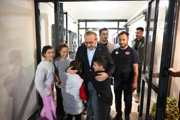 First image shows a man in a suit embracing and interacting with several smiling children in colorful casual clothes near a blue storefront with Aid organization signage and crescent moon logo, surrounded by adults including women in headscarves and men in various attire. Second image depicts the same man in a suit smiling while holding a young girl in a yellow sweater with patterned pants on his arm, with other men in coats visible in the background. Third image captures the man in a suit hugging a boy while a group of children and adults including women in headscarves stand in a hallway lined with bookshelves. Fourth image shows the man in a suit touching the face of a girl in pink while surrounded by children and adults in a corridor setting with some wearing vests featuring a crescent moon emblem.