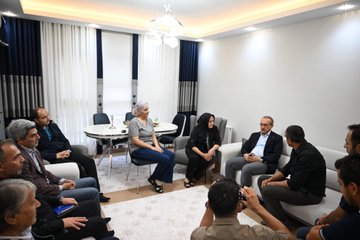 First image shows a man in a blue suit standing near an elevator door in a hallway with wooden panels and decorations like a hanging artwork and shoes on the floor, accompanied by several people including women in headscarves and men in casual attire. Second image depicts three individuals posing with a decorative box, the central man in a blue suit holding it alongside a man in black shirt and a woman in black attire with headscarf, set in a living room with ornate shelves and pillows. Third image captures a group of people seated around a round table in a room with curtains and lighting, engaged in conversation, including men in suits and women in headscarves, with a photographer in the foreground.