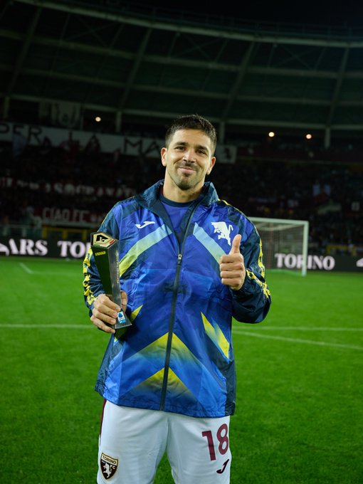 A male soccer player with short dark hair and a beard stands on a green soccer field in front of a goal net wearing a blue and yellow hooded jacket with Torino FC logo white pants with number 18 and black socks holding a tall golden trophy in his right hand raised slightly and making a thumbs up gesture with his left hand the background shows a stadium with banners advertising sponsors like Joma and Torino under stadium lights at night