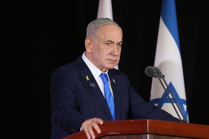 First image: Man in dark suit with blue tie and yellow ribbon pin stands at wooden podium speaking into microphone with Israeli flags on black background. Second image: Group of people including children and adults in casual clothing walk along dirt road through extensive rubble and destroyed multi-story buildings under clear blue sky with scattered clouds.