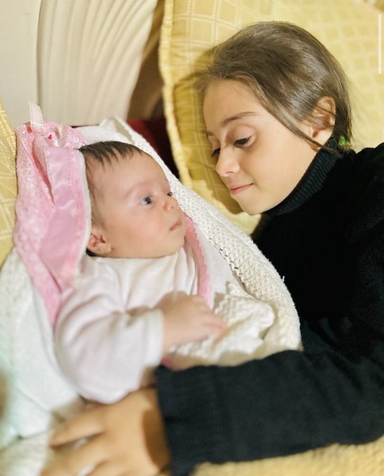 A young girl with brown hair wearing a black sweater sits on a bed with yellow pillows and white decorative elements in the background holding a newborn baby wrapped in a white outfit and pink blanket with a pink bow on its head both looking at each other tenderly