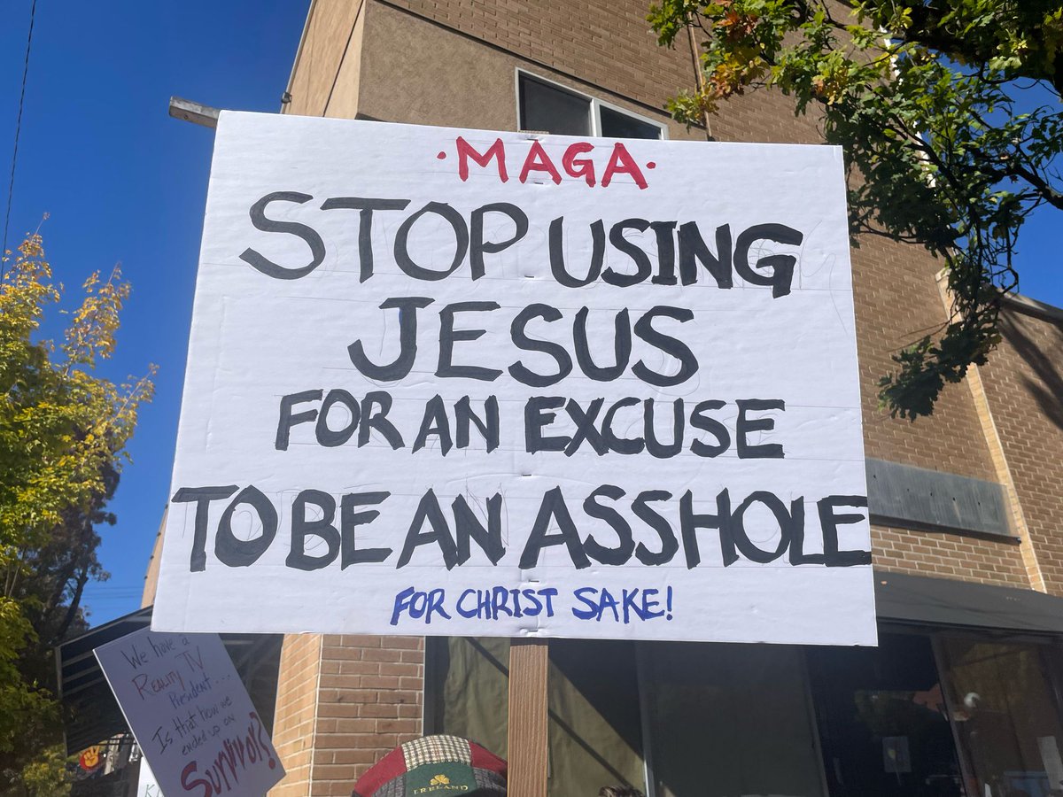 A white protest sign with bold red lettering stating MAGA: Stop using Jesus for an excuse to be an asshole for christ sake! held up in front of a beige brick building under a clear blue sky with green trees nearby and another sign partially visible on the right.