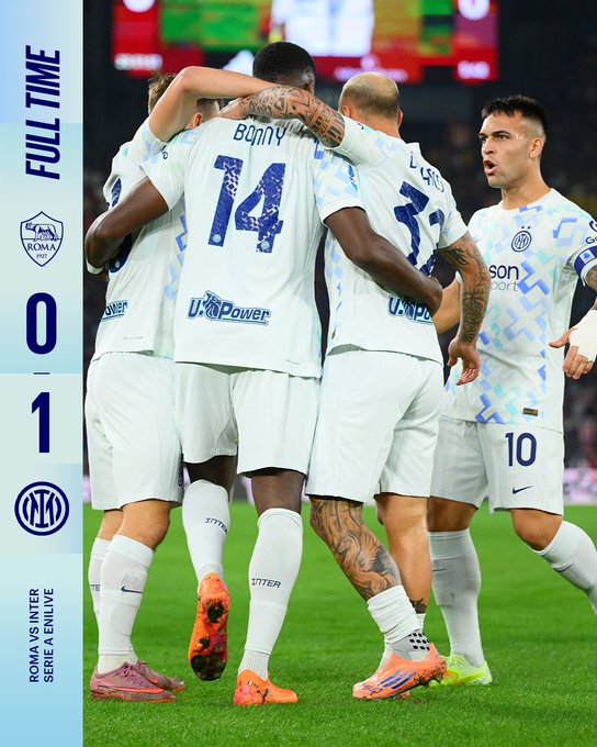 Four soccer players in blue and black Inter Milan uniforms numbered 10, 14, 23, and another embrace in celebration on a green field, with Marcus Thuram identifiable by his jersey, arms linked around shoulders, facing a stadium screen showing full time score Roma 0-1 Inter, team logos visible including ECRA and U-Power sponsors.