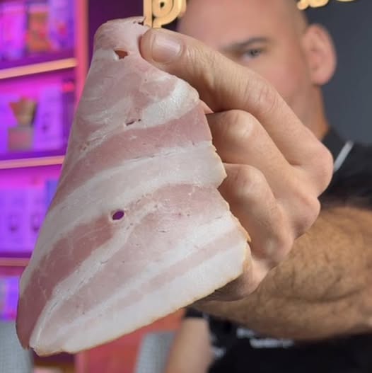 A bald man with blue eyes wearing a black shirt holds a large triangular slice of raw pink ham or pork meat featuring white fat layers marbling and small circular holes in his right hand extended toward the viewer. The background includes a purple-lit kitchen counter with stacked colorful packaged food items and hanging utensils suggesting a food preparation or market setting.