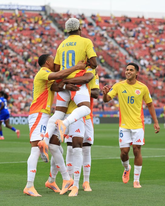 Several male soccer players in yellow jerseys with orange accents and white shorts celebrate a victory by hugging on a green grass field. One player with a white jersey number 10 and another with number 16 stand nearby. The stadium seating in the background is filled with spectators wearing red and other colors. Adidas logos are visible on the jerseys and shorts.