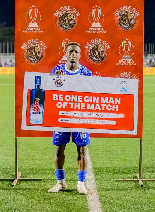 A male soccer player in blue uniform with number 13 stands on a grassy field holding a large check labeled Be One Gin Man of the Match for APR Match X with amount 700,000 crossed out, next to a tall bottle of Be One Gin, surrounded by orange banners featuring lion emblem and Rwanda Premier League text, under stadium lights at night.