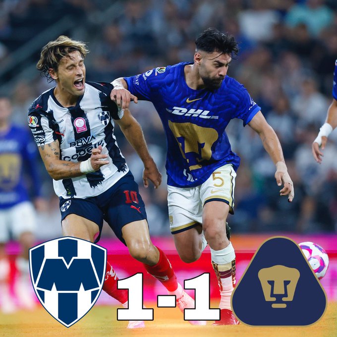 Two soccer players compete for the ball on a stadium field during a match. One player in a white and blue striped jersey with number 10 reaches with his left hand while wearing red shorts and socks. The other player in a blue jersey with number 5 and Puma logo pushes back wearing white shorts and socks. The score 1-1 is displayed at the bottom with team logos of Rayados and Pumas.