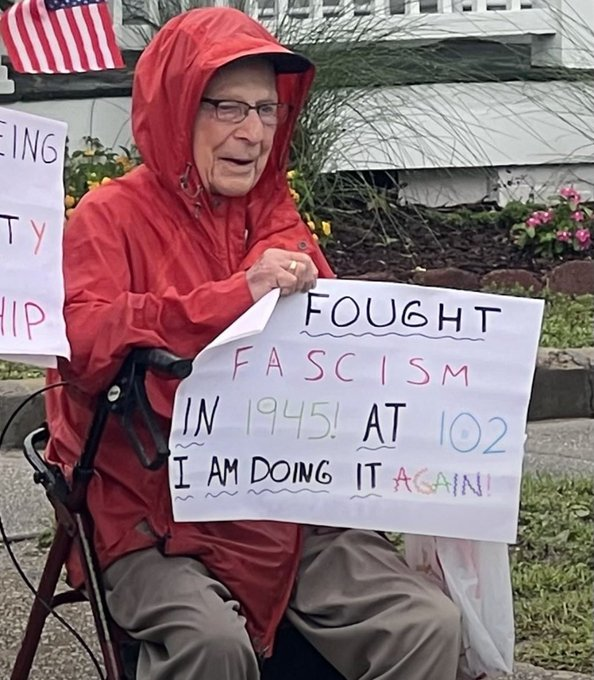 Elderly man with glasses in red hooded raincoat and gray pants sits in wheelchair holding sign that reads I fought fascism in 1945 at 102 I am doing it again. He wears a cap and has a serious expression. American flags wave nearby on porch with white railing and green shrubs. Yellow flowers bloom in garden bed behind. Another sign partially visible says something about fighting fascism.