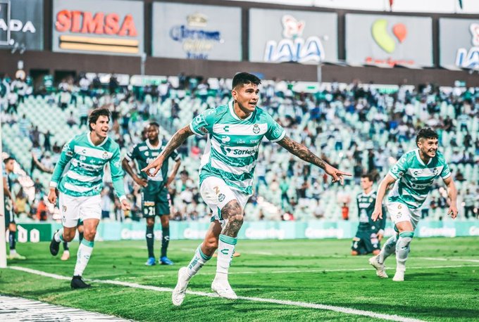 Group of male soccer players in green and white striped jerseys and white shorts, with tattoos visible on some arms, running and celebrating on a green grass field during a match, with a stadium crowd and advertising banners in the background including SMSA and Corona logos.