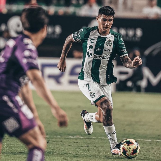A soccer player with dark hair and tattoos on his arms wearing a green and white Santos Laguna jersey with sponsor logos including Soriana dribbles a yellow and colorful soccer ball on a green field during a match against an opponent in a purple jersey with number 2 on the back while another player in green pursues him near the sideline with stadium seating and advertising boards visible in the background.