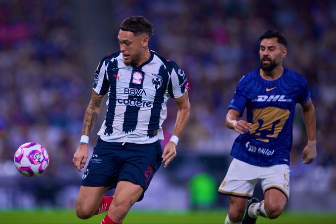 Two soccer players in action on a stadium field during a match one in white and blue striped jersey with pink ball at feet tattooed arms and red shoes the other in blue jersey with white shorts running nearby crowd in background pink sponsor logos visible on jerseys.