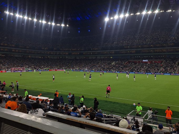 A nighttime soccer stadium filled with spectators under bright lights shows a green field with players in white and blue uniforms positioned during active play near the center circle while referees in orange shirts stand nearby and orange barriers separate the pitch from seated fans in the foreground.