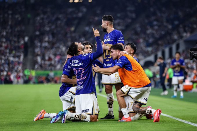Group of male soccer players in purple and white uniforms kneeling and standing on green field, celebrating with raised arms and hugs, one pointing upward, wearing cleats and jerseys with sponsor logos, stadium seating and crowd visible in blurred background under stadium lights.