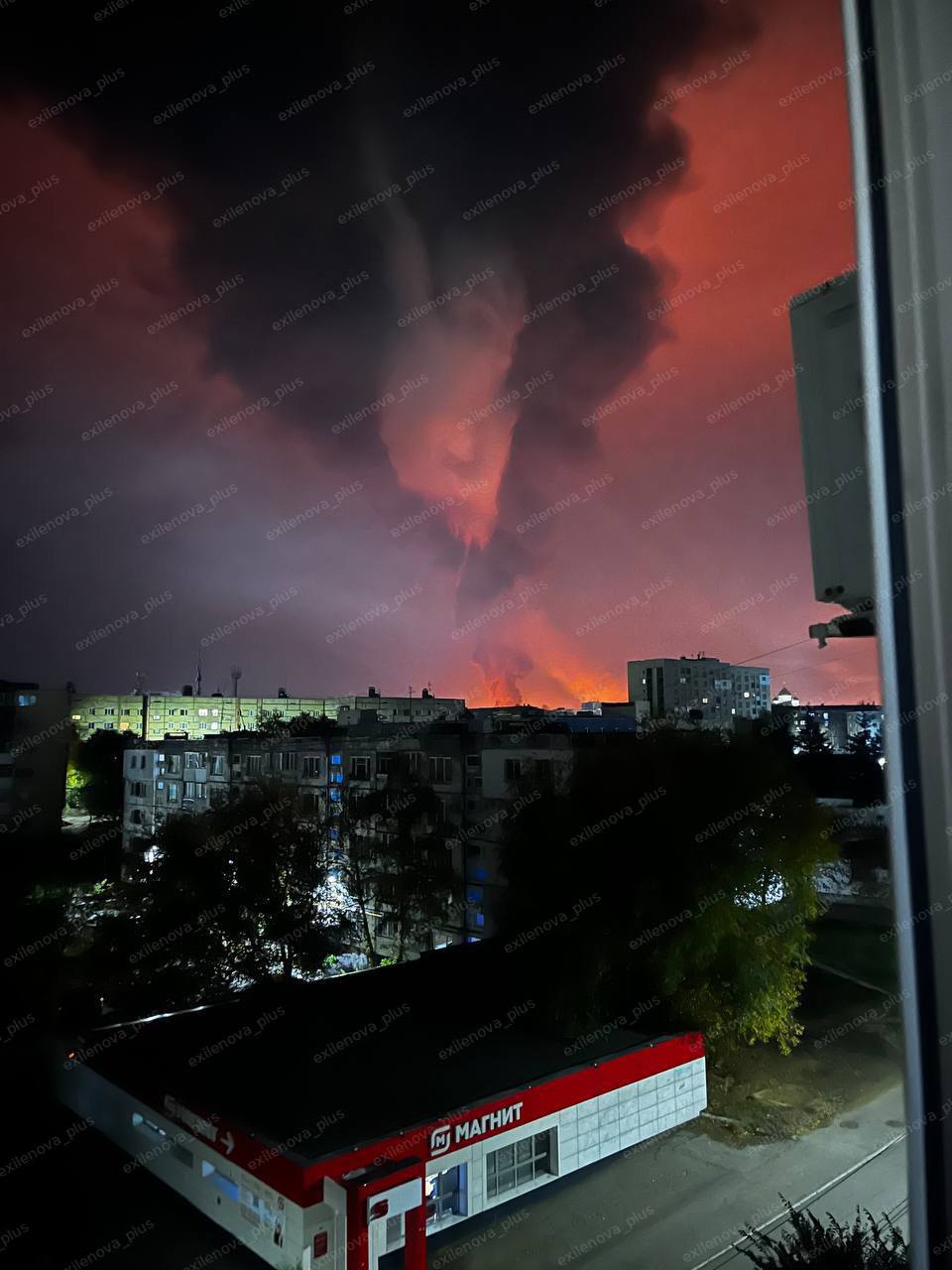 從窗戶看到的夜景顯示，一大股濃稠的黑色和橙色煙霧從一個大型工業設施中滾滾而起，表明烏克蘭無人機襲擊後，薩馬拉地區的新庫伊比雪夫斯克煉油廠發生火災。周圍的城市建築和樹木在燈火通明的天空的映襯下熠熠生輝。在靠近煙霧底部的前景中可以看到一座帶有標牌的紅白相間的建築。