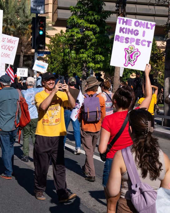 Pedro Pascal stands in a yellow t-shirt and jeans, smiling while holding a phone to take a photo, amid a group of protesters on a street with palm trees and buildings. Protesters wear casual clothes like hats, backpacks, and colorful tops, holding signs reading The only king I respect in purple with a cartoonish figure, Is what crazy like in red, and other banners. An American flag is visible, along with traffic lights showing red and a crosswalk ahead under clear daylight.