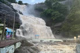 Cascading waterfall flows down rocky cliffs surrounded by dense green trees and mist, with blue safety barriers, railings, steps, and a small building visible at the base amid muddy ground from flooding.