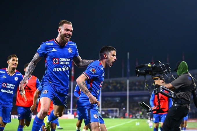 Several male soccer players in blue Cruz Azul jerseys with sponsor logos like Cemex and Novibol celebrate a goal on a nighttime stadium field, one player with tattoos raises his arms in excitement while another smiles broadly, a cameraman in green cap films the scene from the sidelines, additional players in similar uniforms join the celebration near the goalpost.