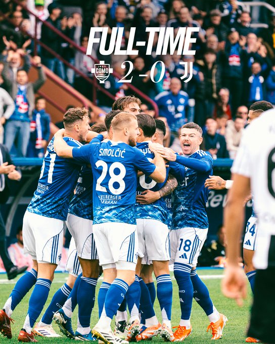 Players in blue and white kits with numbers like 28 and 18 huddle and embrace on green field after match, stadium seating and crowd visible in background, full-time graphic overlay shows Como 2-0.