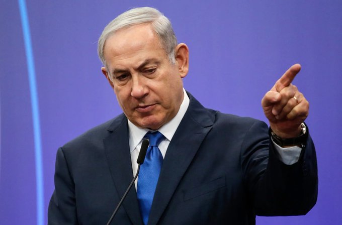 First image shows a man with gray hair wearing a dark suit, white shirt, and blue tie, standing at a podium with a microphone, gesturing with his right hand raised and index finger pointing, against a purple background with a vertical light beam. Second image depicts two Israeli Air Force F-15 fighter jets in formation flight over a sandy desert terrain under a clear blue sky, with one jet slightly ahead and the other trailing, both equipped with external fuel tanks and missiles visible on underwing pylons, and a distant horizon of arid landscape.