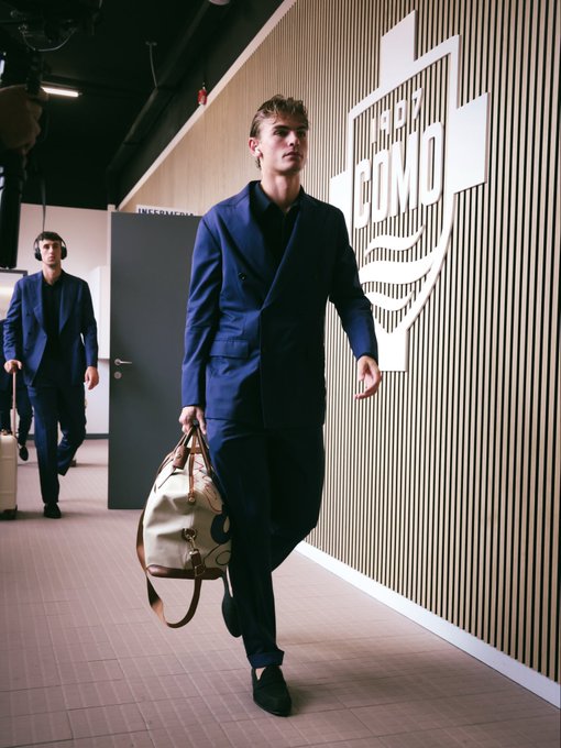 Young man with tousled blond hair dressed in a tailored navy blue double-breasted suit over a white collared shirt walks confidently holding a light brown leather duffel bag featuring a circular emblem. He wears black loafers and strides on a light gray floor toward a dark gray door. Behind him another man in a black suit carrying a small black suitcase follows. The background includes beige wooden slatted walls with white geometric paneling and large white Como 1907 lettering above a wave-like design.