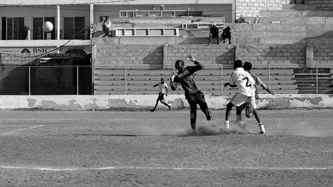 Black-and-white photograph captures a dynamic soccer moment on a dusty field with a goalkeeper in dark uniform extending arms to catch the ball mid-air while two opposing players in white and light jerseys one numbered 7 jump and reach nearby dust kicks up from their feet several other players run in the background blurred stands with spectators and buildings visible behind a fence.