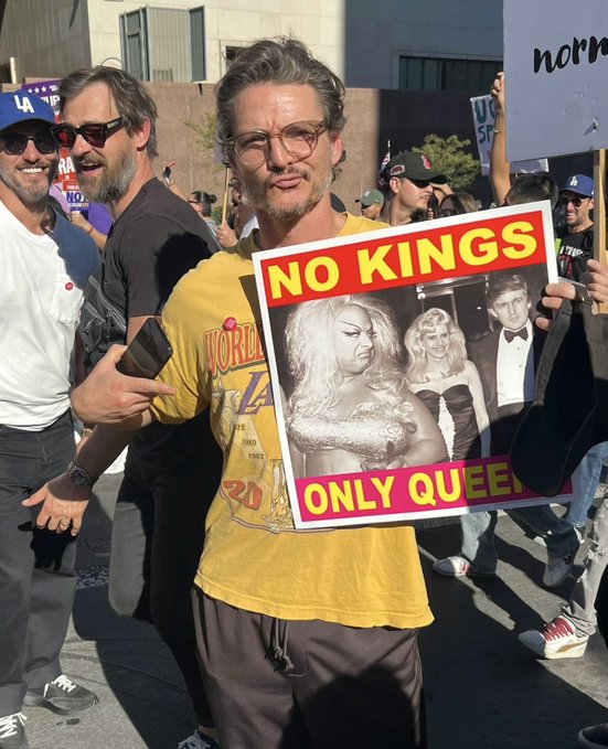 Pedro Pascal stands holding a yellow protest sign reading No Kings Only Queens with satirical collage images of a blonde woman in drag and a man in tuxedo resembling Donald Trump. He wears yellow Los Angeles Lakers t-shirt, gray pants, black cap, and sunglasses, smiling. Group of men in casual clothes and additional protest signs visible in background on sunny urban street.