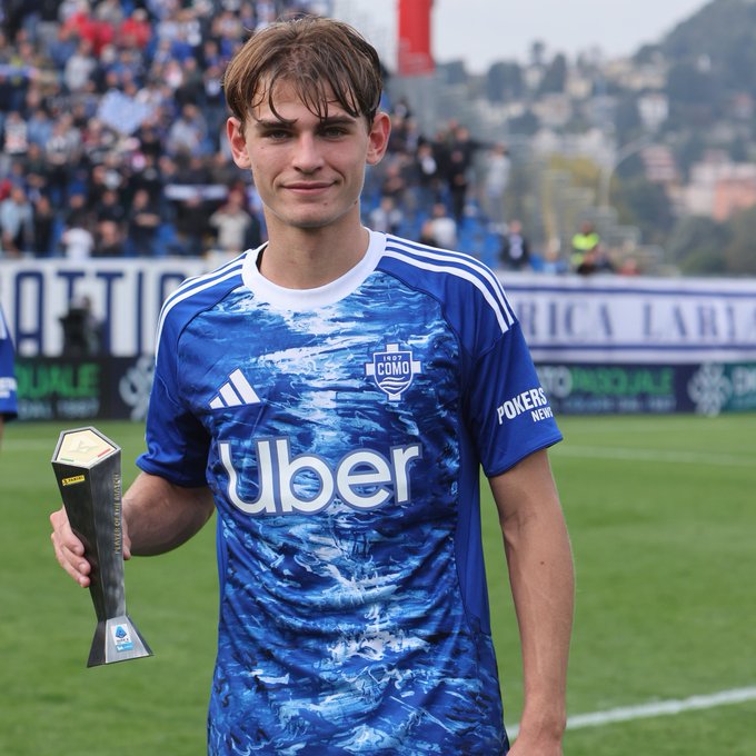 A young male soccer player with short brown hair stands on a green field holding a gold trophy shaped like a shield with a banner. He wears a blue jersey with white wave patterns, Como1907 emblem, Adidas stripes, and Uber sponsor. Stadium seating with spectators and a red banner are in the background, along with a forested hill.