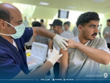 First image shows several men in traditional Saudi attire and casual clothing gathered around a wooden table in a bright indoor hallway with beige walls and glass panels one man in white thobe and red headscarf signs a document while others observe and assist nearby with a TVTC logo visible on the wall. Second image depicts a medical professional in blue scrubs and cap administering an injection to a patient wearing a white thobe in a spacious indoor area with beige walls other individuals in casual wear and medical attire stand around observing the procedure. Third image features a bald man in blue scrubs and mask injecting a young man in gray thobe on the arm in a well-lit room with beige walls and ceiling lights several young men in casual clothing queue nearby with vaccine boxes on a table. Fourth image illustrates a man in blue scrubs and glasses giving an injection to a young man in gray thobe and cap in an indoor setting with beige walls multiple young men in casual attire stand in line watching the process with vaccine supplies on a table.