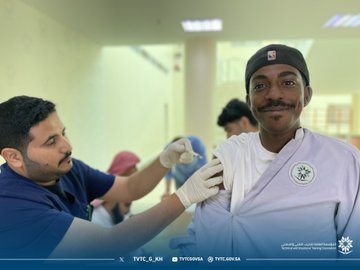 First image shows several men in traditional Saudi attire and casual clothing gathered around a wooden table in a bright indoor hallway with beige walls and glass panels one man in white thobe and red headscarf signs a document while others observe and assist nearby with a TVTC logo visible on the wall. Second image depicts a medical professional in blue scrubs and cap administering an injection to a patient wearing a white thobe in a spacious indoor area with beige walls other individuals in casual wear and medical attire stand around observing the procedure. Third image features a bald man in blue scrubs and mask injecting a young man in gray thobe on the arm in a well-lit room with beige walls and ceiling lights several young men in casual clothing queue nearby with vaccine boxes on a table. Fourth image illustrates a man in blue scrubs and glasses giving an injection to a young man in gray thobe and cap in an indoor setting with beige walls multiple young men in casual attire stand in line watching the process with vaccine supplies on a table.