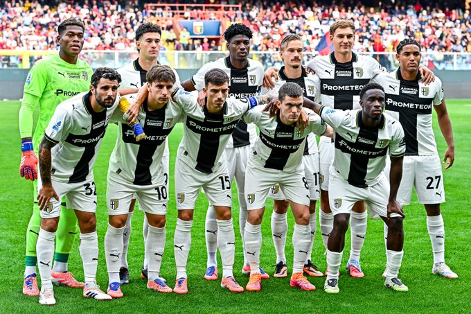 Eleven Parma Calcio players stand in a group pose on a green soccer field with a stadium crowd in the background, wearing white and black striped uniforms with numbers like 15, 10, 27 visible, some with arms around each other, goalkeeper in green kit on the left, all facing the camera.