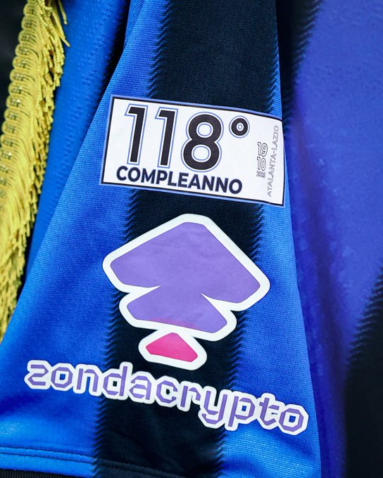 Blue and black striped soccer jersey sleeve with yellow fringe tassels draped over it. White label on blue background reads Compleanno 118 in black text with Atalanta below. Purple abstract arrow-shaped sponsor logo positioned below label. Text Zondacrypto in stylized purple and pink letters at bottom.