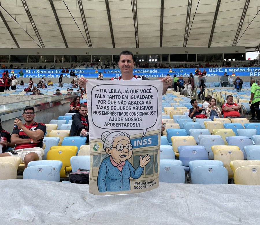 A man in a red Flamengo jersey holds a large yellow banner in a stadium with blue seats and walls. The banner features a cartoon elderly woman in glasses and blue dress gesturing with hand raised, text reading Já que você fala tanto em igualdade por que não abaixa as taxas de juros abusivos nos empréstimos consignados Ajude nossos aposentados, and Não salarial below. Spectators including men and women in casual clothes sit nearby, some watching.
