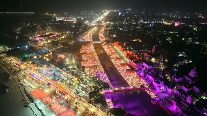 Aerial night view of a long riverbank lined with thousands of small lit oil lamps in colorful patterns forming decorations and pathways. Surrounding area includes illuminated buildings temples and streets with vibrant lights in red orange green and purple hues. The scene captures a festive setup with structured lamp arrangements along the water edge and bridges.