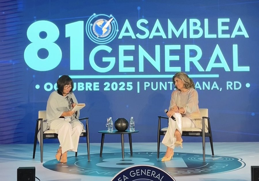 Two women sit on chairs during a panel on a stage with a blue circular backdrop featuring the text 81 ASAMBLEA GENERAL and OEA logo, along with details for the event opening in 2025 in Punta Cana, Dominican Republic. One woman holds a microphone and a notebook, wearing light clothing, while the other holds a microphone and wears light clothing with heels. A small globe sits on a table between them with water bottles.