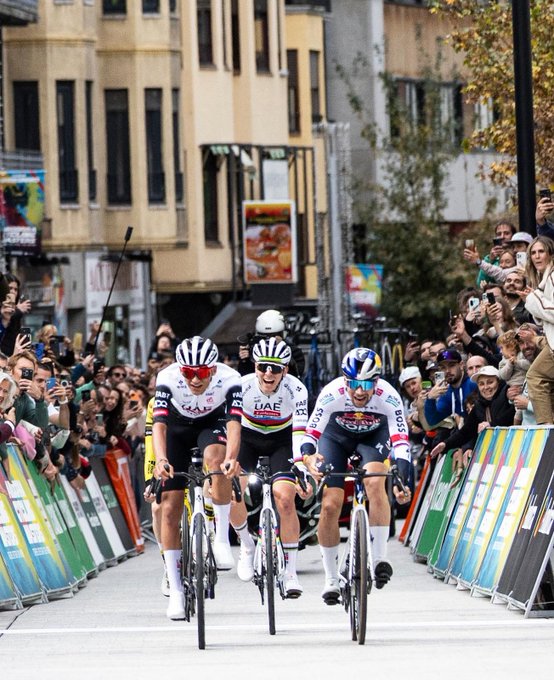 Three cyclists in racing uniforms and helmets cross a finish line on a street lined with barriers and spectators holding phones. The riders wear jerseys from teams like UAE and Red Bull with sponsor logos. Buildings and trees form the urban backdrop under clear skies.