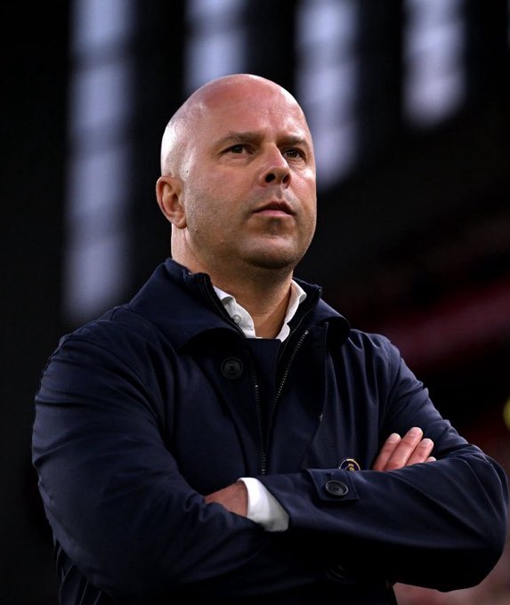 Bald man with serious expression wearing dark jacket over white shirt arms crossed standing in front of dark stadium background with lighting