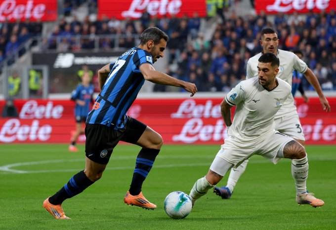 Players in blue and black striped Inter Milan jerseys and white Real Madrid jerseys compete on green soccer field during match. Davide Zappacosta in number 77 blue jersey controls white ball with right foot near sideline. Opponent in white jersey number 18 lunges to challenge. Red and white sponsor banners for Lotto and green stadium seating with crowd in background. Blue team flags wave.