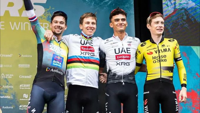 Four male cyclists stand on a colorful podium during the Andorra Cycling Masters event, with Isaac del Toro in second place wearing a UAE Team Emirates jersey, Primož Roglič in first with a rainbow-striped champion jersey, and two others in XRG and Visma-Lease a Bike uniforms, arms raised in celebration against a blue background with sponsor logos.