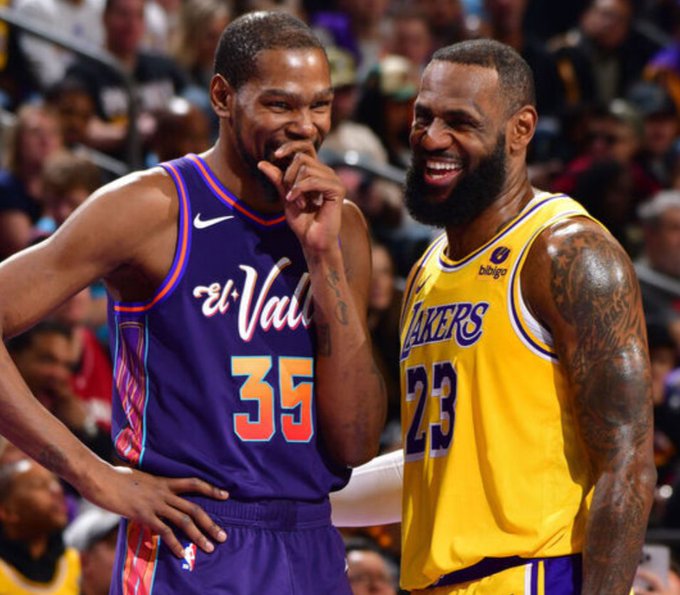 Kevin Durant in purple Phoenix Suns jersey number 35 stands next to LeBron James in yellow Los Angeles Lakers jersey number 23, both smiling with hands near mouths in a basketball arena crowd setting.