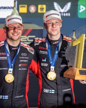Two male drivers in red and black racing suits stand on a podium holding gold medals and a trophy, smiling with arms around each other; one wears a white Hankook cap. Background includes sponsor banners for Denes, CCI, Panasonic, Tamdac, and Toyota team logos. Another image shows a black Hyundai rally car numbered 8 parked on grass amid yellow autumn trees and a crowd in the distance. A third image focuses on the rear of a black rally car with sponsor logos like Panasonic Automoto and CCI on a dirt path surrounded by orange and yellow fall leaves and trees.